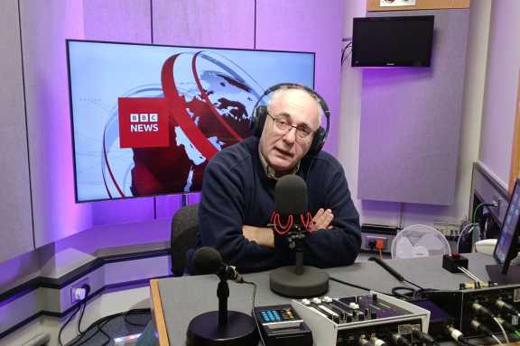 Dave recording at the BBC sitting in front of mic with BBC logo in the background 